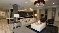 This single-floor space showcases a refined modern design with an open-concept layout that integrates the living and kitchen areas into one cohesive, elegant environment. The living area is anchored by a plush light-gray sofa adorned with geometric red and white accent pillows, paired with a clean-lined white coffee table that grounds the space. A striking drum chandelier with crystal detailing serves as a bold focal point overhead, casting a warm, ambient glow throughout. Recessed lighting complements the overall illumination, highlighting the thoughtful layering of light in the space. The kitchen features dark subway tile backsplash, dark cabinetry with under-cabinet lighting, and a sleek countertop that extends into a bar-style seating area — ideal for casual dining or entertaining. Charcoal and dark wood tones in the kitchen contrast beautifully with the lighter palette of the living area, creating a dynamic yet harmonious visual flow. A leather accent chair and a lush potted plant add textural variety and a touch of organic warmth to the space. Rich dark flooring ties the entire layout together, contributing to the space's sophisticated, contemporary ambiance.