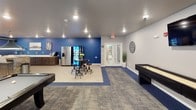 This single-floor amenity space is thoughtfully designed to serve as a social hub, combining a comfortable lounge area with an active game room. The lounge is anchored by a striking floor-to-ceiling brick fireplace with a wall-mounted television above, surrounded by plush leather seating arrangements and patterned carpeting over blue-bordered flooring. Multiple windows fitted with white blinds allow natural light to fill the space, complemented by recessed ceiling lighting throughout.

Adjacent to the lounge, a wet bar area features a marble-topped counter with white bar stools, pendant lighting, and a bold blue accent wall—ideal for entertaining. The game room section is well-equipped with a pool table, shuffleboard table, dartboard, and arcade cabinet, along with a wall-mounted TV and decorative artwork, creating an energetic atmosphere for recreation.

A separate area provides additional communal space with high-top tables, industrial-style stools, a vending machine, and a stainless steel refrigerator set against a vibrant blue accent wall. French doors lead to connected areas, and consistent patterned carpet with blue border detailing ties all zones together cohesively.