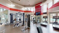 This fitness center is designed to deliver a comprehensive workout experience within a spacious, single-floor layout. The room's most striking architectural feature is its soaring two-story ceiling, lined with a row of clerestory windows that flood the interior with natural light and lend an open, airy feel. Bold red horizontal stripes along the upper walls add a dynamic, energetic aesthetic that complements the facility's purpose.

The main workout area is generously proportioned, accommodating a diverse range of equipment without feeling crowded. Cardio options include multiple treadmills, stationary upright and recumbent bikes, and elliptical machines, all positioned to take advantage of the surrounding views through large windows and glass doors that open toward an outdoor pool area. Strength training is well-represented with a selection of cable and resistance machines, a dedicated free weights section with a dumbbell rack, and additional functional training stations.

Large floor-to-ceiling mirrors along one wall visually expand the space and allow users to monitor their form during workouts. The flooring throughout is a durable, textured rubber surface suited to heavy use. Pendant lighting hangs from the ceiling, supplementing the natural light. An adjacent entry corridor, framed by glass doors with tile flooring, provides access to a computer workstation area, suggesting this facility is part of a broader amenity complex.
