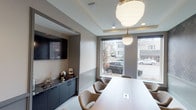 This refined single-floor commercial space is designed to impress, offering a polished and professional environment ideal for meetings and collaboration. The centerpiece of the space is a sleek conference room, accessible through dramatic full-glass double doors framed in matte black, providing a striking first impression while offering a clear sightline into the interior.

Inside, the conference room features a long, smooth conference table surrounded by upholstered seating in warm neutral tones. Overhead, a pair of elegant globe pendant lights with gold accents hang from a subtly recessed tray ceiling, lending the room a refined, contemporary character. Recessed lighting complements the ambient glow throughout.

A built-in media wall with dark cabinetry and granite-topped surfaces houses a large flat-screen display, providing both functional and aesthetic value. Decorative accents, including a globe and framed artwork, add personality without overwhelming the professional atmosphere.

Large windows along the exterior wall invite abundant natural light and offer views of the surrounding streetscape, creating an open, connected feel. The flooring is a standout feature — a bold geometric pattern combining light wood tones with dark tile creates a visually dynamic foundation that ties the space together. Neutral warm-gray walls and dark trim details throughout contribute to the space's cohesive, upscale aesthetic.