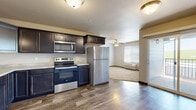 Located at 3148 Champion Dr in Rapid City, South Dakota, this single-floor residence offers a well-organized layout designed for comfortable everyday living. The kitchen serves as a functional centerpiece, outfitted with dark espresso-toned cabinetry featuring raised-panel doors and light countertops. Stainless steel appliances — including a refrigerator, electric range with microwave above, and dishwasher — provide a cohesive, modern aesthetic. A pendant light fixture and wood-style plank flooring complete the kitchen's polished look. The kitchen opens directly into the living area, which transitions to plush carpeting and features a ceiling fan with light kit, a wall-mounted HVAC unit, and a window fitted with blinds that allows natural light to filter in. A glass-paned exterior door offers additional access and light. The bedroom areas are carpeted and neutrally finished, with one room featuring a walk-in closet with built-in shelving. The bathroom includes a full soaking tub and shower combination, tile-style flooring, and a dark vanity with a light countertop. Throughout the unit, dark trim accents provide a consistent design detail that ties the interior together.