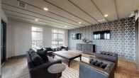 Located at 4015 Aspen Grove Drive in Franklin, Tennessee, this stylish single-floor lounge space is designed with a refined, contemporary aesthetic that blends comfort and sophistication. The main gathering area is anchored by warm hardwood flooring laid in a herringbone-influenced pattern, complemented by a striking geometric wallcovering that serves as a bold focal point. Mounted flat-screen televisions and a substantial herringbone-front media credenza enhance the entertainment-focused layout. The ceiling features a distinctive linear paneled design with recessed lighting that adds architectural depth and a warm, inviting ambiance. Generously sized windows along one wall flood the interior with natural light and frame views of the surrounding Franklin cityscape. Tufted leather seating, a large rustic wood coffee table, and layered area rugs contribute to the space's upscale yet approachable character. A well-appointed wet bar area with subway tile backsplash, gray cabinetry, under-counter refrigeration, and a stainless steel sink provides excellent functionality for entertaining. On the opposite side of the room, a set of French doors opens directly onto a spacious outdoor terrace, where additional seating areas and sweeping views of the Tennessee hills create an exceptional extension of the interior living space.