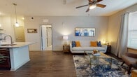 This single-floor residence at San Mateo Crossing offers a thoughtfully designed open-concept layout that seamlessly connects the living, dining, and kitchen areas. The living room features rich hardwood-style flooring, elevated ceilings with a ceiling fan, and neutral-toned walls that create a bright, welcoming atmosphere. A large area rug and glass-top coffee table anchor the space, while recessed lighting and wall art add refined detail.

The kitchen is a standout feature, centered around a generous granite-topped island with an undermount sink and bar seating. Rich dark wood cabinetry extends to the ceiling, providing ample storage, while stainless steel appliances — including a refrigerator, range, and microwave — complete the modern aesthetic. Pendant lighting above the island adds warmth and style.

The bedrooms offer plush carpeted flooring and natural light through blinds-fitted windows. One bedroom includes a walk-in closet, while both are well-proportioned and serene. The bathroom is finished with large-format tile flooring, a glass-enclosed shower, dark wood vanity with a marble-style countertop, and contemporary gold-accented fixtures. The entry features durable wood-look flooring and a paneled front door.