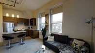 This single-floor space showcases an open-concept design that seamlessly integrates the living and kitchen areas into one cohesive layout. The living area is furnished with a sleek black leather sofa, accented by a glass coffee table and soft textiles, creating a comfortable yet modern atmosphere. Two large windows flood the space with natural light while offering views of the surrounding urban environment, enhancing the sense of openness. The kitchen is outfitted with rich, dark wood cabinetry that provides ample storage, complemented by light stone countertops and stainless steel appliances. A breakfast bar with seating offers a casual dining option that connects the kitchen to the living space. The flooring throughout features a polished concrete surface, lending an industrial character that ties the design together. Neutral wall tones keep the space feeling bright and airy, while decorative touches such as a tall indoor plant and urban-themed wall art add personality. Track lighting installed along the ceiling provides functional and adjustable illumination. The overall ambiance balances contemporary urban style with practical, comfortable living.