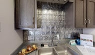 Located at 9100 Andrews Hwy in Odessa, Texas, this single-story residential unit offers a practical and well-appointed living space designed for comfortable everyday living. The layout is efficiently organized, with distinct areas flowing cohesively throughout the floor plan.

The living area is bright and inviting, featuring durable wood-look plank flooring and a textured ceiling. A large sliding glass door fitted with vertical blinds allows generous natural light to pour in, creating a warm and airy atmosphere. An adjacent room, finished with contrasting gray-toned walls and a similarly styled window, provides a versatile secondary space ideal for a bedroom or home office.

The kitchen is a functional highlight of the unit, showcasing dark espresso-toned cabinetry paired with gray granite-look countertops. A standout decorative metallic tile backsplash adds a distinctive, textured focal point behind the double-basin stainless steel sink. The kitchen also accommodates a full-size refrigerator and additional appliances, offering ample storage and prep space.

The bathroom is clean and efficiently designed, featuring a single-sink vanity with a granite-look countertop and dark wood cabinetry, a large mirror, and a full tub-shower combination with white tile surround. The space is finished in neutral tones throughout, contributing to a cohesive and move-in-ready presentation.