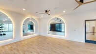 Located at 7243 US-431 N in Owens Cross Roads, Alabama, this single-floor fitness and dance studio is thoughtfully designed to provide an open, energizing environment for a variety of movement-based activities. The studio's most striking architectural features are its large arched mirrors framed with built-in LED backlighting, creating a dramatic yet functional focal point that enhances both the sense of space and the studio's modern aesthetic. Light-toned hardwood flooring runs throughout, offering a clean, resilient surface well-suited for exercise, dance, or group fitness classes. The space is bathed in natural light through expansive floor-to-ceiling windows dressed with sheer curtains, offering views of the surrounding area while maintaining an airy, welcoming atmosphere. Modern ceiling fans with sleek, multi-blade designs are positioned throughout to ensure consistent airflow and comfort. Recessed lighting supplements the natural light, providing even illumination across the open floor plan. A dedicated weight and equipment area accommodates strength training, while the open studio floor provides ample room for group classes or personal training sessions. Glass-paneled entry doors and clearly marked exits contribute to the space's clean, professional design. Overall, the studio presents a polished and functional environment designed to support an active, health-focused community.