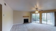 This single-floor residence centers around a comfortable living area designed with both function and ease in mind. The main living space is finished with plush wall-to-wall carpeting in a neutral grey tone, complementing the warm beige walls that lend the room a calm, inviting atmosphere. A wood-manteled fireplace serves as a natural focal point, adding a classic, cozy element to the space. Above, a ceiling fan with an integrated light fixture provides both air circulation and ambient lighting, contributing to the room's practical design. Natural light flows generously into the space through a full-width sliding glass door, which opens directly onto a private balcony enclosed by a sleek metal railing. The balcony overlooks a lush, tree-lined outdoor setting, bringing a pleasant sense of greenery and tranquility to the interior. The sliding door arrangement also creates a seamless visual connection between the interior living area and the outdoor space. The overall aesthetic is clean and straightforward, with simple architectural lines and a neutral color palette that offers a versatile backdrop adaptable to a variety of furnishing styles.