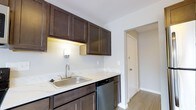 This single-floor space presents a clean, modern aesthetic with thoughtful design choices throughout. The kitchen serves as a focal point, showcasing rich dark-toned wood cabinetry that provides ample upper and lower storage. Light stone-patterned countertops offer a striking contrast against the darker cabinetry, while a stainless steel sink, dishwasher, and full-size refrigerator complete the suite of appliances. Under-cabinet lighting adds both functionality and a warm ambiance to the workspace. The kitchen's layout is efficient and well-organized, allowing for comfortable use of the space. Light wood-tone flooring runs throughout, contributing to a bright and cohesive feel. The crisp white walls enhance the sense of openness and allow natural light to reflect freely across the space. A doorway visible from the kitchen leads further into the unit, suggesting a practical and connected floor plan. Overall, the space strikes a balance between modern finishes and functional design, making it a comfortable and visually appealing environment.