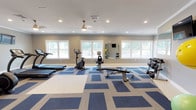This fitness center offers a spacious, well-appointed environment designed to support a comprehensive workout routine. The room is finished with neutral gray walls, white crown molding, and a distinctive blue-and-tan geometric patterned carpet that adds a modern aesthetic to the space. Recessed lighting throughout, complemented by ceiling fans and pendant fixtures, ensures the area remains well-lit and comfortable during any workout.

Large white-trimmed windows line the walls, flooding the space with natural light and offering pleasant outdoor views. A full-length wall mirror enhances the sense of openness while allowing users to monitor their form during exercise.

The cardio section is equipped with multiple treadmills and a stationary upright bike, all from SportsArt, as well as a rowing machine and recumbent bike. The strength training area features a tiered dumbbell rack with a full range of weights, kettlebells, and a weight bench. Additional functional training equipment includes resistance bands, exercise balls, and foam rollers, neatly stored on a dedicated wall-mounted rack. A wall-mounted flat-screen television is also present for entertainment. The fitness center connects to an adjacent space, providing convenient access to other amenities.