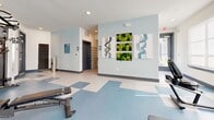 Located at 3842 W Northwest Highway in Dallas, TX, this fitness center offers a thoughtfully designed workout facility across a single floor. The exercise room is the heart of the space, featuring a blue and white checkered vinyl floor that adds a clean, modern aesthetic. Large wall-mounted mirrors visually expand the room while allowing members to monitor their form during workouts. Natural light filters in through multiple windows, creating a bright and energizing atmosphere, complemented by recessed lighting and a ceiling fan overhead. The cardio area is equipped with multiple treadmills and an upright bike, while a full dumbbell rack with a range of free weights and colorful medicine balls supports strength training. A multi-station cable machine anchors one end of the room for versatile resistance exercises, alongside an adjustable workout bench. The walls are painted in a soft, calming blue-gray tone and adorned with decorative wall art featuring moss accents and abstract designs, adding a refreshing visual element to the space. A connected entryway leads to an accessible restroom, finished with coordinating tile flooring and clean, minimal décor. Posted fitness policies near the entrance ensure a well-managed environment for all users.