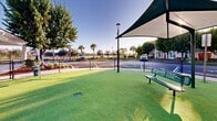Located at Everblades Parkway North in Estero, Florida, this generously sized outdoor dog park offers a well-maintained and thoughtfully designed space for pets and their owners. The ground is covered entirely in vibrant artificial turf, providing a clean, comfortable surface year-round regardless of Florida's variable weather conditions. Expansive shade sail canopies are strategically positioned throughout the area, offering relief from the Florida sun while keeping the space open and airy. Green metal benches are placed beneath the shaded areas, giving owners a comfortable place to relax while supervising their pets. Agility elements, including tunnel accessories, are integrated into the space to encourage active play and engagement. The entire area is enclosed by a secure chain-link fence, ensuring a safe environment for off-leash activity. A covered wash station structure sits at one end of the park, providing a convenient area for post-play pet care. The surrounding landscape features mature trees, palm trees, and manicured shrubs that add natural beauty and further shade to the environment. The park enjoys clear sightlines and an open sky, making it a bright, welcoming amenity set within a vibrant residential community.