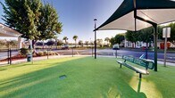 Located at Everblades Parkway North in Estero, Florida, this generously sized outdoor dog park offers a well-maintained and thoughtfully designed space for pets and their owners. The ground is covered entirely in vibrant artificial turf, providing a clean, comfortable surface year-round regardless of Florida's variable weather conditions. Expansive shade sail canopies are strategically positioned throughout the area, offering relief from the Florida sun while keeping the space open and airy. Green metal benches are placed beneath the shaded areas, giving owners a comfortable place to relax while supervising their pets. Agility elements, including tunnel accessories, are integrated into the space to encourage active play and engagement. The entire area is enclosed by a secure chain-link fence, ensuring a safe environment for off-leash activity. A covered wash station structure sits at one end of the park, providing a convenient area for post-play pet care. The surrounding landscape features mature trees, palm trees, and manicured shrubs that add natural beauty and further shade to the environment. The park enjoys clear sightlines and an open sky, making it a bright, welcoming amenity set within a vibrant residential community.