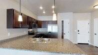 This single-floor space showcases a well-appointed kitchen that serves as its centerpiece. The kitchen is defined by rich, dark espresso-toned cabinetry that extends to the upper walls, providing generous storage while creating a bold visual contrast against the light neutral walls and ceiling. The countertops are finished in speckled granite, offering both durability and an elegant aesthetic that carries through to the expansive kitchen island, which features a double stainless steel sink and serves as an ideal space for meal preparation or casual dining.

Stainless steel appliances, including a French door refrigerator with an ice and water dispenser, a range with an electric cooktop, and an over-the-range microwave, complete the kitchen's modern and cohesive look. Recessed lighting is complemented by a sleek pendant light suspended above the island, providing focused task lighting with a contemporary touch.

The layout flows openly, with vaulted or raised ceilings that enhance the sense of spaciousness. A doorway visible in the background hints at additional rooms or storage beyond, suggesting a practical and thoughtfully designed floor plan throughout the space.