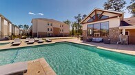 Located at 2400 N Morrison Blvd in Hammond, Louisiana, this outdoor amenity area serves as a centerpiece of a well-appointed residential community. The space is anchored by a generously sized swimming pool with crystal-clear water, complemented by a separate spa pool featuring a decorative water fountain. In-water lounge chairs are integrated directly into the shallow end of the pool, offering a resort-style relaxation experience. The expansive concrete pool deck is lined with numerous cushioned chaise lounges, providing ample space for sunbathing beneath the open Louisiana sky. A standout architectural feature is the craftsman-style clubhouse structure adjacent to the pool, characterized by prominent exposed wood beam gables, stone column accents, and floor-to-ceiling glass windows that flood the interior with natural light. A covered outdoor lounge area with comfortable seating extends from the clubhouse, creating a shaded retreat. Additional outdoor seating and gathering areas are scattered throughout the deck. The community's multi-story residential buildings frame the backdrop, surrounded by mature pine trees that provide a natural, wooded setting. The entire area is enclosed by decorative metal fencing, ensuring privacy and security while maintaining an open, airy atmosphere throughout the complex.