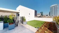 This expansive rooftop terrace offers a thoughtfully designed outdoor retreat with a clean, modern aesthetic defined by crisp white parapet walls and large-format tile flooring throughout. The space is divided into several functional zones, each contributing to an inviting and versatile environment. A dedicated lawn area features lush artificial turf bordered by mulched planting beds, providing a green focal point against the urban backdrop. Generous landscaping throughout includes ornamental trees in bold planters, native-style succulents, ornamental grasses, and shrub plantings that add texture and color. An outdoor dining area is furnished with a large table and chairs, illuminated by overhead string lights that create a warm ambiance during evening gatherings. Adjacent to the dining area, a built-in BBQ grill station is set beneath a covered pergola structure, complete with counter space and planter-integrated cabinetry. A dedicated lounge zone features plush rounded sectional seating and accent chairs, perfectly positioned to take in panoramic city skyline views. The surrounding urban skyline, visible from all corners of the terrace, provides a dramatic and ever-present backdrop, making this rooftop an exceptional space for both relaxation and entertainment.