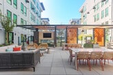 Located at 2230 Calle de Luna in Santa Clara, CA, this generously sized single-floor amenity space offers a sophisticated blend of modern design and versatile functionality. The interior lounge area immediately impresses with its layered ceiling detail—geometric wood-trimmed coffers set against crisp white surfaces—creating an architectural focal point that draws the eye upward. Bold terracotta-red velvet seating anchors the lounge, complemented by a built-in wood-paneled nook with integrated shelving, offering an inviting alcove for relaxed gatherings. A large-format wall-mounted display and striking abstract artwork add curated character throughout.

The adjacent dining and catering kitchen area is finished with warm wood cabinetry, a marble-effect backsplash, and a sleek central dining table surrounded by teal velvet chairs. Banquette seating lines one wall, bathed in natural light that streams through expansive windows. Clean-lined tile flooring runs throughout, maintaining a bright and cohesive aesthetic.

Black-framed glass double doors connect the interior to a spacious outdoor terrace, where pergola structures with warm-toned wood slats provide shade over lounge and dining furniture. An outdoor fireplace and mounted TV make the terrace a true extension of the entertaining space, all set against the backdrop of the surrounding modern residential building.