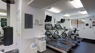 Located at 500 Garden Ln in Huntington, West Virginia, this fitness center offers a focused and functional workout environment designed to meet everyday exercise needs. The single-floor space is equipped with multiple treadmills, including StairMaster Pro models, arranged to maximize the use of the available floor space. Wall-to-wall mirrors line one side of the room, creating a sense of expanded space while allowing users to monitor their form during workouts. Each treadmill station features its own mounted flat-screen TV, adding convenience and entertainment during cardio sessions. The room is well-lit with overhead fluorescent lighting that ensures a bright and energizing atmosphere. Practical amenities are thoughtfully integrated throughout, including a small utility shelf stocked with cleaning spray for equipment sanitation, a dehumidifier to maintain air quality and comfort, and a waste bin for easy disposal. An electrical panel is accessible on the wall, and a wall-mounted clock keeps exercisers on schedule. A window on one end of the room allows for a touch of natural light. The space maintains a clean, white-walled aesthetic with dark flooring, contributing to an orderly and motivating fitness environment.