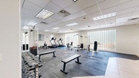 Located at 380 Brazilian Circle in Port Saint Lucie, Florida, this dedicated fitness center offers a generously sized, single-floor exercise room designed to support a full range of workout routines. The space features a drop-panel ceiling with recessed fluorescent lighting that evenly illuminates the entire floor, creating a bright and energizing atmosphere. A striking glass block wall allows diffused natural light to filter into the room, adding an open, airy feel without compromising privacy.

The exercise floor is covered with durable rubber matting, providing both safety and comfort underfoot. A full-length mirrored wall expands the visual depth of the space and allows users to monitor their form during workouts. Equipment is thoughtfully arranged throughout the room and includes treadmills, a rowing machine, a cable cross machine, a weight-assisted strength station, flat and adjustable benches, and a comprehensive dumbbell rack with Rogue flat benches. A stability ball and exercise mat round out the functional training options.

The neutral-toned walls and clean finishes contribute to a professional yet welcoming environment. A bulletin board area near the entrance provides a practical organizational touch. French doors lead to an adjacent area with light-toned flooring, offering additional transitional space. Overall, this fitness center is well-suited for a community or facility setting.
