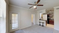 This 1-bedroom, 1-bathroom single-level unit offers a thoughtfully arranged floor plan that maximizes everyday comfort and functionality. The open-concept living area features plush carpeting, crown molding, and a ceiling fan with integrated lighting, creating a bright and welcoming atmosphere. A large window with blinds allows natural light to fill the space while offering privacy. The living area flows seamlessly into the kitchen, which is defined by a raised breakfast bar with bar stool seating — ideal for casual dining or entertaining. The kitchen features dark countertops that contrast with lighter cabinetry, and a full-size refrigerator is visible, indicating ample storage and appliance space. A hallway visible from the main living area leads to the private bedroom and bathroom, providing a clear separation between living and sleeping spaces. The neutral color palette throughout — from the light gray walls to the soft carpeting — lends the unit a clean, versatile aesthetic that adapts easily to a variety of décor styles. Crown molding and consistent trim details add a refined architectural touch to this functional and comfortable single-floor residence.