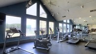 This fitness center offers an energizing workout environment defined by its striking architectural design and thoughtfully curated equipment. The space is anchored by soaring vaulted ceilings that create an open, airy atmosphere, complemented by a dramatic arrangement of large, floor-to-ceiling windows that flood the interior with abundant natural light. Deep blue accent walls provide a bold, modern contrast to the bright white ceiling, lending the space a sleek and motivating aesthetic. A ceiling fan and track lighting ensure year-round comfort and optimal visibility throughout the facility. The workout floor is outfitted with a comprehensive selection of cardio equipment, including stationary bikes, elliptical trainers, treadmills, and stair climbers, all arranged to maximize space and airflow. Large mirrors line one wall, enhancing the sense of spaciousness while allowing users to monitor their form during exercise. Expansive windows on multiple sides offer views of the surrounding outdoor areas, including a fenced terrace and neighboring buildings, keeping the space connected to its environment. The durable flooring is well-suited for high-traffic fitness use, and the overall layout promotes ease of movement between machines. This fitness center is a functional and visually impressive space designed to support a complete cardio workout experience.