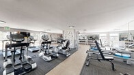 Located at 240 S Monaco St Pkwy in Denver, CO, this fitness center offers a generously sized, single-floor exercise room designed to support a comprehensive workout experience. The space is thoughtfully laid out, with distinct zones for cardio and strength training, separated by a decorative lattice-style partition that adds an architectural touch to the interior.

The cardio section features a robust lineup of True-brand treadmills, elliptical machines, stationary bikes, and WaterRower rowing machines, all positioned on dark carpet tiles that contrast with the light-toned large-format tile flooring throughout the rest of the space. Floor-to-ceiling mirrors line portions of the walls, enhancing the sense of openness and allowing users to monitor their form.

The strength training area is equally well-appointed, with a full dumbbell rack, a cable machine, and multiple weight-stack resistance machines. An accent wall featuring a bold navy and yellow horizontal stripe adds a vibrant, energetic character to the space, reinforcing its athletic purpose.

Natural light filters in through windows along the perimeter, offering views of the surrounding outdoor landscape. Wall-mounted televisions provide entertainment, and the clean, neutral color palette — complemented by bright flush-mount lighting — creates an inviting, functional environment suited for a wide range of fitness routines.