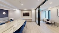 Located at 35 David Salomon Drive in Toronto, this single-floor communal space offers a thoughtfully designed environment suited for both social gatherings and focused meetings. The layout is divided into distinct functional zones, seamlessly connected by a hallway and anchored by a central living area.

The main lounge area features a built-in wood-panelled media wall with a mounted flat-screen display, complemented by a large oval marble-top dining table surrounded by navy blue upholstered chairs. A lounge nook along one wall is defined by a bold navy accent wall adorned with two large-scale abstract artworks in warm earth and blue tones. A long built-in grey bench sofa runs the length of this nook, paired with contemporary marble-top pedestal tables and rounded amber accent stools, creating a casual and inviting atmosphere.

Black-framed glass partition walls divide the space into semi-private meeting areas, each equipped with a compact round table and chairs, wall-mounted air conditioning units, and access to natural light through floor-to-ceiling windows. Light wood-look flooring flows throughout, unifying the space with a clean, modern aesthetic. Recessed lighting overhead ensures a bright, welcoming ambiance, while curated artwork and thoughtful furnishings lend the space a polished, professional character.