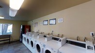 This single-floor laundry facility is designed for practical, efficient use. The main area features a continuous row of front-loading washing machines paired with matching dryers, all aligned along a neutral beige wall, creating a clean and organized layout. The machines are commercial-grade units, suggesting a shared or community-use setting. Informational notices and warning signs are mounted on the wall above the machines, keeping users informed of rules and operating instructions. A Coca-Cola vending machine is positioned at one end of the room, offering a convenient amenity for users during their visit. The flooring appears to be durable tile, well-suited for a high-traffic laundry environment. Recessed lighting runs along the ceiling, providing adequate illumination throughout the space. The overall color palette—beige walls, white machines, and neutral flooring—gives the room a clean, utilitarian appearance. The layout is straightforward and accessible, with ample space along the front of the machines for loading and unloading laundry comfortably.
