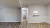 This single-floor unit offers a clean, updated interior with a cohesive design throughout. The living area features wood-look flooring and a step-up platform section, adding a subtle architectural detail to the open layout. French doors at the far end of the living space allow natural light to filter in and provide an inviting focal point. A decorative arched pass-through connects the living area to the kitchen, which is well-equipped with white shaker-style cabinetry, granite countertops, a full suite of white appliances including a dishwasher, electric range, built-in microwave, and refrigerator. The kitchen also benefits from wood-look tile flooring that complements the rest of the common areas. A hallway leads to the private quarters, where the bedrooms are finished with plush carpeting and fitted with window blinds that offer outdoor views and light control. The primary bedroom includes a ceiling fan with lighting and multiple windows, enhancing ventilation and brightness. The bathroom features a tiled shower-tub combination, wood-look flooring, and access to a dedicated closet space. Throughout the unit, neutral wall tones and consistent finishes create a bright, move-in-ready atmosphere.
