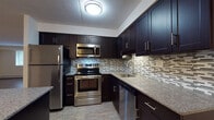 This single-floor space offers a well-designed layout that combines modern finishes with functional living. The kitchen is a standout feature, showcasing rich dark cabinetry with sleek hardware, granite countertops, and a full suite of stainless steel appliances including a refrigerator, electric range, built-in microwave, and dishwasher. A mosaic tile backsplash in neutral tones adds a refined decorative element, while a center island with a matching granite surface provides additional prep space and casual seating. Light wood-style flooring runs throughout, creating a warm, cohesive aesthetic. The open living area is generously proportioned, offering flexible arrangement possibilities. Natural light filters in through windows that frame views of surrounding greenery, and a ceiling fan provides additional comfort. Baseboard heating lines the perimeter of the living space. The bathroom is finished with large-format tile on both the floor and tub surround, complemented by a dark vanity with granite countertop and brushed nickel fixtures, maintaining the consistent design language found throughout the unit.