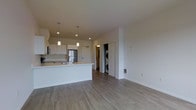 This single-floor residence showcases a clean, modern design with an open-concept layout that seamlessly connects the kitchen and living areas. The kitchen is outfitted with white shaker-style cabinetry, stainless steel appliances including a full-size refrigerator, and a peninsula island with an integrated sink that doubles as a breakfast bar. Pendant lighting hangs above the island, adding a stylish focal point to the space. Recessed lighting throughout ensures the area remains bright and well-illuminated. Durable wood-look luxury vinyl plank flooring runs consistently across the entire floor, lending a warm yet contemporary feel to the space. The open living area offers a generous, versatile footprint ideal for a variety of furniture arrangements. A dedicated in-unit laundry closet houses a stacked washer and dryer, providing added convenience. Additional doorways suggest access to a bedroom and a walk-in closet or storage area. The overall palette of crisp white walls, neutral flooring, and modern fixtures creates a bright, airy ambiance that feels both functional and inviting.