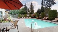 This inviting outdoor amenity space centers around a generously sized swimming pool with clear, turquoise water, accessible via stainless steel entry rails and surrounded by a textured concrete pool deck. A row of sling-style lounge chairs lines the perimeter of the pool, providing ample space for sunbathing and relaxation. Multiple red market umbrellas offer shaded retreats throughout the space, including over a cast-iron patio dining table and chairs, ideal for outdoor meals or casual gatherings. Vibrant seasonal flower arrangements in large decorative planters add bursts of color throughout the area, enhancing the welcoming atmosphere. The space is framed by neatly trimmed columnar hedges and a perimeter fence, creating a sense of privacy and enclosure. Towering evergreen trees rise above the boundary, providing a lush green backdrop and natural windbreak. Adjacent to the pool deck, a building with large multi-pane windows and neutral siding serves as a clubhouse or amenity facility, its interior visible through the glass. The overall ambiance is bright, well-maintained, and well-suited for outdoor leisure, combining functional poolside amenities with thoughtfully curated landscaping for a pleasant and resort-like experience.