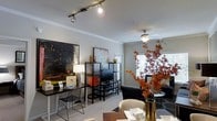 This single-floor residence showcases a thoughtfully designed open-concept layout that seamlessly blends multiple functional areas into one cohesive living space. The interior is characterized by neutral-toned walls accented with crown molding, contributing to an elegant and polished aesthetic throughout.

The living area is anchored by a ceiling fan and bathed in natural light streaming through shuttered windows, creating a bright and airy atmosphere. A plush sofa and coordinating side table furnish the space comfortably, while decorative branches in a tall vase add a striking organic accent.

Adjacent to the living area, a media zone features a sleek black metal shelving unit housing a flat-screen television, complemented by decorative objects and storage boxes in warm accent tones. Bold abstract artwork adorns the walls, lending an artistic character to the space.

A dedicated workspace is incorporated into the layout, complete with a dark writing desk, a stylish wire-frame chair, and a table lamp, offering a functional yet design-forward area for work or study.

The dining area, positioned in the foreground, is set with tableware and surrounded by upholstered seating, making it ideal for entertaining. Track lighting overhead provides focused illumination, enhancing the modern, urban feel of this well-appointed residence.
