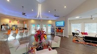 This single-floor community clubhouse is designed to impress, combining striking architectural details with a lively, welcoming atmosphere. The main lounge commands attention with soaring vaulted ceilings accented by ambient blue uplighting and a sculptural pendant light fixture, while ceiling fans and recessed lighting ensure comfort throughout. Wide-plank hardwood-style flooring runs seamlessly across the space, grounding the vibrant décor.

The central gathering area features modern white lounge chairs arranged around a glass-top table adorned with a bold floral arrangement, set atop a vivid multicolored area rug that energizes the room. An adjacent lounge zone offers a large white sectional sofa with red accent pillows, additional seating, and abundant natural light pouring through multiple large windows dressed in white curtains and blinds.

A business center station with orange bar-height seating and a wall-mounted flat-screen TV provides a functional workspace, while French doors along one wall open to an outdoor deck, seamlessly connecting the interior to the outdoors. The kitchen features granite countertops, white cabinetry, stainless steel appliances, and vivid red pendant lights above an eat-in bar with orange seating — a cheerful space suited for entertaining and resident gatherings alike.