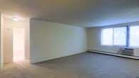 This single-floor space presents a straightforward and functional layout centered around a generously sized open room. The interior is finished with wall-to-wall carpeting in a neutral gray tone, providing a soft underfoot feel that complements the clean, understated aesthetic throughout. The walls are painted in a light, creamy hue, contributing to a bright and airy atmosphere even in the absence of abundant natural light sources.

A single window positioned along one wall allows natural light to filter into the space, casting a soft glow that enhances the room's openness. The window appears to be a standard unit with a simple frame, providing both ventilation and a connection to the exterior. Beneath the window runs a baseboard heating unit, a practical feature that ensures efficient climate control along the perimeter of the room.

The ceiling is smooth and standard in height, maintaining a clean, unadorned appearance consistent with the rest of the space. An electrical outlet is visible along the lower portion of one wall, indicating convenient access for furnishings and appliances. Overall, the space offers a blank canvas with a calm, neutral ambiance well-suited for a variety of uses and personal customization.