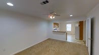 This single-floor residence offers a straightforward, functional layout designed for comfortable everyday living. The main living area is finished with wall-to-wall carpeting, providing a warm and soft underfoot feel, while crisp white walls create a bright, clean backdrop throughout the space. A ceiling fan with integrated lighting fixture is centrally mounted, ensuring consistent air circulation and overhead illumination. Recessed lighting complements the ceiling fan, adding to the overall brightness of the room.

The living area opens seamlessly toward the kitchen, which is connected via a raised breakfast bar with a countertop overhang — ideal for casual dining or additional workspace. The kitchen features warm wood-toned cabinetry, a white range, and a full-size refrigerator, offering practical storage and cooking essentials. The kitchen floor transitions to a classic checkered tile pattern, visually distinguishing it from the carpeted living space. A window in the kitchen area allows natural light to flow in, enhancing the open feel of the combined living and kitchen zone.

The space benefits from a well-maintained HVAC system with ceiling vents providing climate control. The clean, neutral palette and open arrangement make this a versatile and move-in ready residence.