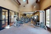 Located at 5082 N Primitivo Way in Fresno, California, this fitness center offers a dedicated exercise room designed to support a comprehensive workout experience. The space is defined by its striking architectural character, featuring soaring vaulted ceilings with exposed dark wood beams that lend a warm, Southwest-inspired aesthetic. Warm-toned walls and carpet flooring create an inviting atmosphere throughout.

The exercise room is generously outfitted with a wide variety of equipment to accommodate both cardio and strength training needs. Cardio machines include treadmills, elliptical trainers, and stationary bikes, while the strength training area features multi-station cable machines, free weight benches, and a full dumbbell rack. Floor-to-ceiling mirrors along one wall visually expand the space and allow users to monitor their form during workouts. A wall-mounted flat-screen television provides entertainment during cardio sessions.

Natural light filters in through high clerestory windows and large glass entry doors, which offer views of the surrounding landscaped outdoor area. Recessed lighting supplements the natural light, ensuring the space is well-lit throughout the day. The glass-paneled entry doors provide convenient access directly from the exterior, creating a seamless connection between indoors and the outdoors.