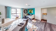 This contemporary two-floor residence showcases a thoughtfully designed interior that blends industrial character with modern comfort. The main living area commands attention with dramatically soaring double-height ceilings, floor-to-ceiling windows, and an abundance of natural light that pours in from multiple exposures, offering views of the surrounding urban landscape. Exposed concrete ceilings and textured grey walls lend an industrial-loft aesthetic, while wide-plank hardwood flooring runs throughout, unifying the space.

The open-plan living and dining area is anchored by statement pendant lighting and large multi-pane windows, creating a bright and airy atmosphere. A well-appointed kitchen features stainless steel appliances including a gas range, a decorative mosaic tile backsplash, white quartz countertops, and a mix of wood-toned and high-gloss cabinetry.

The primary bedroom offers a serene retreat with a ceiling fan, walk-in closet with built-in shelving, and an en suite bathroom finished with a frameless glass shower, wood-grain vanity, LED-backlit mirror, and a bold green accent wall. A second full bathroom mirrors this quality with a soaking tub, matching vanity, and teal accent wall. A secondary sleeping area with direct balcony access and vibrant teal accent wall completes this well-appointed residence.