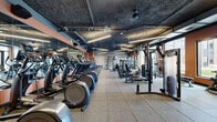 Located at 2650 University Ave SE in Minneapolis, MN, this fitness center offers a comprehensive and thoughtfully designed workout environment across a generously sized single floor. The facility is divided into two distinct zones, each with its own aesthetic and purpose. The main strength and cardio area features an industrial-style design with exposed ceilings, visible ductwork, and linear lighting that creates an energetic atmosphere. Rubber-tiled flooring supports rows of treadmills, ellipticals, stair climbers, and a wide array of strength machines and cable systems. Warm terracotta-toned accent walls and large windows allow natural light to fill the space and provide outdoor views, while floor-to-ceiling mirrors amplify the sense of openness. A dedicated entry area includes cubbies, a water station, and recycling bins for convenience. The second zone contrasts with a bright, modern aesthetic featuring light hardwood-style flooring, white walls, and a drop ceiling with recessed lighting. This studio-style space is equipped with rowing machines, spin bikes with display screens, a ballet barre, kettlebells, medicine balls, freestanding boxing bags, and open floor space ideal for functional and group training. A textured stone accent wall with a mounted display screen adds a refined finishing touch to this versatile training area.