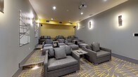 This stylish private screening room is designed with both comfort and aesthetics in mind. The space is anchored by a large wall-mounted projection screen paired with a ceiling-mounted projector, creating a true cinematic experience. Seating is arranged in a tiered layout, with generously sized gray upholstered lounge chairs and sofas, each accompanied by small side tables for added convenience. The flooring features a striking patterned carpet in gray and yellow, which complements the room's bold color palette. Walls are finished in a cool gray tone, accented by a vibrant yellow textured accent wall near the screen that adds a pop of energy to the space. Sleek vertical wall sconces provide warm, ambient lighting throughout, while recessed ceiling lights offer additional illumination. Abstract artwork adorns the side walls, contributing to the room's contemporary character. A glass door at the rear of the space allows natural light to filter in and provides a view of an outdoor area. The overall ambiance is sophisticated and inviting, blending modern design elements with a relaxed, lounge-like atmosphere well-suited for private screenings or social gatherings.