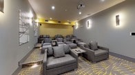 This stylish private screening room is designed with both comfort and aesthetics in mind. The space is anchored by a large wall-mounted projection screen paired with a ceiling-mounted projector, creating a true cinematic experience. Seating is arranged in a tiered layout, with generously sized gray upholstered lounge chairs and sofas, each accompanied by small side tables for added convenience. The flooring features a striking patterned carpet in gray and yellow, which complements the room's bold color palette. Walls are finished in a cool gray tone, accented by a vibrant yellow textured accent wall near the screen that adds a pop of energy to the space. Sleek vertical wall sconces provide warm, ambient lighting throughout, while recessed ceiling lights offer additional illumination. Abstract artwork adorns the side walls, contributing to the room's contemporary character. A glass door at the rear of the space allows natural light to filter in and provides a view of an outdoor area. The overall ambiance is sophisticated and inviting, blending modern design elements with a relaxed, lounge-like atmosphere well-suited for private screenings or social gatherings.