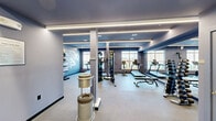 The fitness center at The Cornerstone Westbury, located at 461 Railroad Ave in Westbury, NY, offers a well-appointed workout facility designed to support a comprehensive exercise routine. The space is finished in a cool blue-grey color palette, lending a modern and energizing ambiance throughout. Linear LED lighting runs along the ceiling perimeter, providing bright, even illumination that complements the natural light streaming in through large windows along one wall, which also offer views of the surrounding exterior. Full-length wall mirrors on one side visually expand the space and assist with form during workouts. The cardio zone features treadmills and a stationary cycling bike, positioned directly in front of the windows. A multi-functional cable machine serves as the centerpiece for strength and resistance training, accompanied by a dedicated free weights area complete with a tiered dumbbell rack and an adjustable workout bench. Wall-mounted storage shelving holds stability balls, medicine balls, and rolled exercise mats, ensuring the space stays organized and versatile. The flooring is covered in durable, low-pile carpet suited for fitness use. Structural columns are integrated cleanly into the layout, and access doors lead to adjacent utility or amenity spaces. Overall, the fitness center is thoughtfully equipped and designed for residents seeking a convenient, full-featured workout environment.