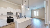 Located at 124 Iberville St in Madison, Alabama, this single-floor unit offers a well-appointed open-concept layout that seamlessly connects the kitchen and living room. The kitchen is a standout feature, showcasing crisp white shaker cabinetry, a large central island with a white quartz countertop and a matte black farmhouse-style faucet, stainless steel appliances, and stylish pendant lighting hanging from the ceiling. Crown molding adds a refined architectural touch throughout the space. The living area is bright and welcoming, with a ceiling fan, a glass-paneled entry door, and windows that invite natural light and outdoor views. Warm hardwood-style flooring runs continuously across the entire unit, lending a cohesive and polished look to every room. The bedroom is generously sized, featuring a ceiling fan, a large window with exterior views, and clean neutral walls that create a calm, restful atmosphere. A well-organized walk-in closet with built-in shelving and hanging rods provides ample storage. The overall aesthetic is modern and clean, with a neutral color palette of soft whites and warm beiges that enhances the sense of openness and light throughout.
