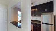 This single-story space in Gallery, US, offers a clean and functional layout with thoughtfully chosen finishes. The kitchen is a standout area, showcasing rich dark espresso-toned shaker-style cabinetry paired with dark granite countertops that feature subtle speckled detailing. Track lighting overhead provides focused illumination, while a pass-through counter connects the kitchen to an adjacent area, enhancing the sense of openness and flow between spaces. Stainless steel appliances complement the kitchen's refined aesthetic.

The adjoining living area benefits from natural light streaming through slatted window blinds, offering a bright and welcoming atmosphere. A separate room, finished with warm light hardwood flooring, features a large window that allows ample daylight to fill the space, creating an airy and versatile environment suitable for a variety of uses. The room's neutral white walls and clean lines contribute to a bright, minimalist feel.

Throughout the space, white trim and molding provide a crisp contrast against the neutral palette, lending a polished and well-maintained appearance. The overall ambiance is modern yet approachable, combining practical design elements with clean finishes.