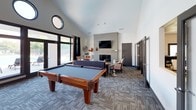This contemporary single-floor community amenity space is designed for both relaxation and recreation. The main gathering area commands attention with dramatic vaulted ceilings that amplify the sense of openness, complemented by a pair of distinctive circular porthole windows that add architectural character near the roofline. Large floor-to-ceiling glass doors and wide windows flood the interior with natural light while offering views of the outdoor patio area beyond.

A full-size billiards table serves as a central recreational feature, positioned on patterned carpet tiles that lend a modern, textural quality to the flooring throughout. The lounge area is anchored by a painted brick fireplace with a wall-mounted flat-screen television above the mantle, creating an inviting focal point. Comfortable navy blue sofas and accent chairs in bold chevron patterns are arranged around a white coffee table to encourage social gathering.

Additional high-top bar seating near the windows provides a casual spot for conversation or casual work. A glimpse into an adjacent room reveals a kitchenette and a separate workspace or meeting area, extending the functionality of this versatile community hub. The overall aesthetic blends clean white walls with dark-trimmed doors and windows, striking a polished yet welcoming balance.