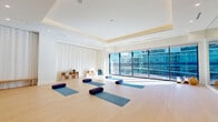 Located at 632 Newark Avenue in Jersey City, NJ, this yoga studio offers a thoughtfully designed, single-floor space that promotes calm and focus. The open studio floor is finished with light hardwood flooring that lends warmth and a sense of spaciousness throughout the room.

One of the studio's most striking architectural features is the wall of floor-to-ceiling windows, which floods the interior with abundant natural light and offers views of the surrounding urban streetscape. Sheer white curtains line the perimeter walls, softening the atmosphere and providing flexible privacy options during classes.

A coffered ceiling with recessed lighting adds a refined architectural detail overhead, while a full-length mirror wall on one side of the studio enhances both the sense of depth and functionality for practitioners monitoring their form. Open-grid wooden shelving units along one wall provide organized storage for props such as bolsters and blocks.

The studio is equipped with yoga mats and cylindrical bolsters arranged across the floor, accommodating group sessions comfortably. A smart panel near the entrance allows for environmental control, while a glass-paneled entry door provides a welcoming, professional threshold. The overall ambiance is clean, minimal, and tranquil — a well-appointed urban wellness space designed to support mindful movement and practice.