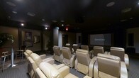 This dedicated home theater is designed to deliver a true cinematic experience within a private setting. The room is finished with a dark ceiling and carefully controlled ambient lighting, creating an immersive atmosphere that minimizes distractions and enhances on-screen visuals. Rows of plush, cream-colored leather recliner seats are arranged in a tiered layout, ensuring unobstructed sightlines from every position in the room. The seating arrangement comfortably accommodates a generous number of guests, making it ideal for both intimate screenings and larger gatherings. At the front of the room, a large projection screen takes center stage, powered by a Denon audio-visual system that provides rich, high-fidelity sound to complement the visual presentation. Recessed ceiling speakers are integrated throughout the space to deliver a full surround-sound experience. The entryway features a clearly marked exit and access door, while subtle wall-mounted artwork adds a refined touch to the otherwise utilitarian design. The overall ambiance is that of a professionally appointed screening room, balancing comfort, functionality, and audiovisual excellence in a single, thoughtfully designed space.