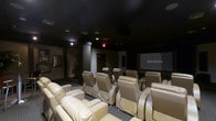 This dedicated home theater is designed to deliver a true cinematic experience within a private setting. The room is finished with a dark ceiling and carefully controlled ambient lighting, creating an immersive atmosphere that minimizes distractions and enhances on-screen visuals. Rows of plush, cream-colored leather recliner seats are arranged in a tiered layout, ensuring unobstructed sightlines from every position in the room. The seating arrangement comfortably accommodates a generous number of guests, making it ideal for both intimate screenings and larger gatherings. At the front of the room, a large projection screen takes center stage, powered by a Denon audio-visual system that provides rich, high-fidelity sound to complement the visual presentation. Recessed ceiling speakers are integrated throughout the space to deliver a full surround-sound experience. The entryway features a clearly marked exit and access door, while subtle wall-mounted artwork adds a refined touch to the otherwise utilitarian design. The overall ambiance is that of a professionally appointed screening room, balancing comfort, functionality, and audiovisual excellence in a single, thoughtfully designed space.