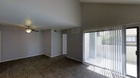 This single-floor space offers a clean, open layout defined by striking architectural details and modern finishes. The living area immediately draws attention with its soaring vaulted ceilings, which create an expansive, airy atmosphere. Dark hardwood-style flooring runs throughout, providing a rich contrast against the neutral gray walls and crisp white trim. A ceiling fan is mounted overhead, adding both functionality and a subtle design element to the space.

Natural light is a standout feature, pouring in generously through a large sliding glass door fitted with vertical blinds, which also provides direct access to an outdoor area. The abundance of sunlight enhances the open feel of the room and highlights the quality of the flooring and finishes.

The space is equipped with multiple HVAC vents, ensuring consistent climate control throughout. A closet door is visible along the back wall, offering practical built-in storage. The overall ambiance is modern and understated, with a neutral color palette that provides a versatile backdrop suitable for a variety of interior styles. The combination of high ceilings, natural light, and quality flooring makes this a bright, welcoming, and highly functional space.