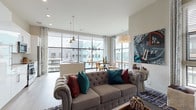 Located in Atlanta, GA 30318, this single-floor residence showcases a thoughtfully designed open layout with light wood-tone flooring running throughout. The main living area is anchored by expansive floor-to-ceiling windows that flood the space with natural light and offer views of the surrounding cityscape. A separate media or sitting room provides additional flexible living space with elevated ceilings.

The kitchen is appointed with high-gloss white cabinetry, a marble-style tile backsplash, stainless steel appliances, and a generous island with bar seating and pendant lighting above—ideal for both cooking and casual dining. Large windows continue in this space, maintaining the bright and airy atmosphere.

The bedroom features light wood flooring, ample natural light, and direct access to a well-appointed en-suite bathroom. The en-suite includes a wide vanity with white cabinetry, a backlit mirror, and a full tub-shower combination with dark tile accents. An additional full bathroom with similar white vanity finishes and light wood flooring serves the rest of the unit. Throughout, the design balances modern minimalism with eclectic, artistic touches.