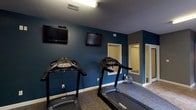 The Reserve's fitness center is a thoughtfully designed, single-floor facility organized across multiple interconnected rooms, providing residents with a comprehensive space for a full-body workout. The main cardio area features multiple treadmills positioned beneath wall-mounted flat-screen televisions, making it easy to stay entertained during endurance training. Bold teal accent walls contrast with neutral gray tones throughout, giving the space a modern, energetic atmosphere complemented by durable carpet flooring suited for athletic use.

A dedicated room houses an elliptical cross-trainer alongside a wall-mounted accessory rack, with natural light filtering in through window blinds to create a bright, welcoming environment. An adjacent strength training room provides open floor space with a weight bench and free weights, offering ample room for resistance and functional fitness exercises. This area also benefits from natural light through a window and features the same consistent gray color palette and carpet flooring found throughout the facility.

A tiled hallway connects the various workout areas, providing smooth transitions between zones and leading to additional amenities visible along the corridor. The overall layout is practical and efficient, ensuring users can move seamlessly between cardio, strength, and open training areas within one cohesive fitness facility.